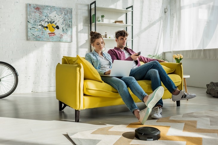 best robot vacuums save you time - couple relaxing on couch while robot vacuum cleans floor